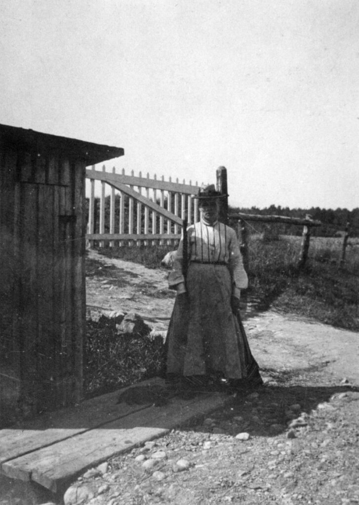 Svartvitt fotografi, kvinna står bredvid liten bod, i bakgrunden grind vid järnvägsövergång.