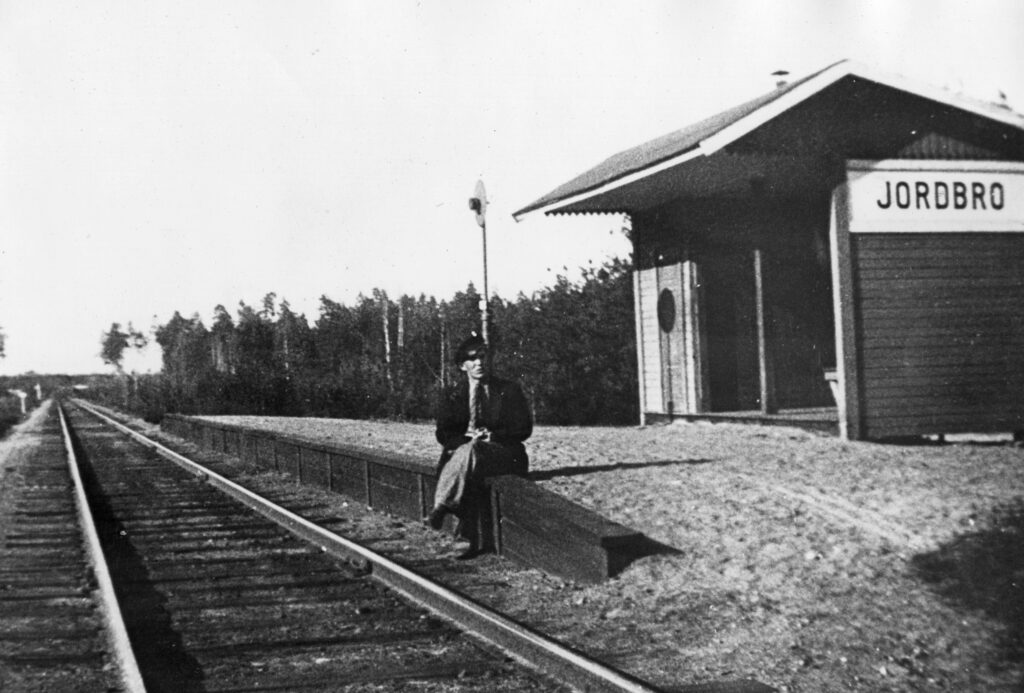 Svartvitt fotografi. En man sitter på gammal perrong med fötterna bredvid rälsen. I bakgrunden stationens väntkur.