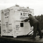 Svartvitt fotografi, man står på trädäck och flyttar en stor vit kontainer märkt med svart text.