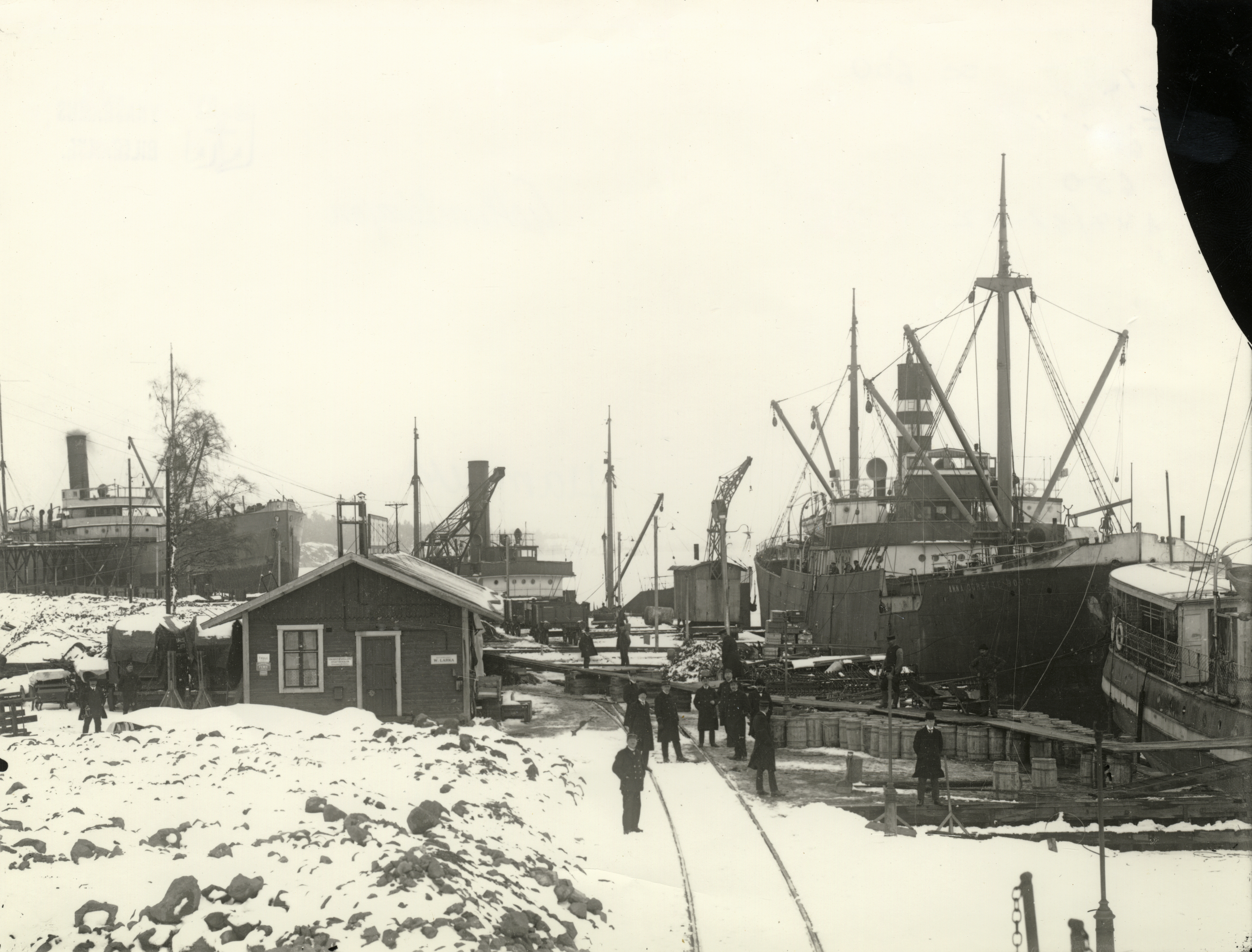 Svartvitt fotografi, på bilden syns förråd och smalspårig järnväg som löper ner till kajen där män lastar malm på fartyg.