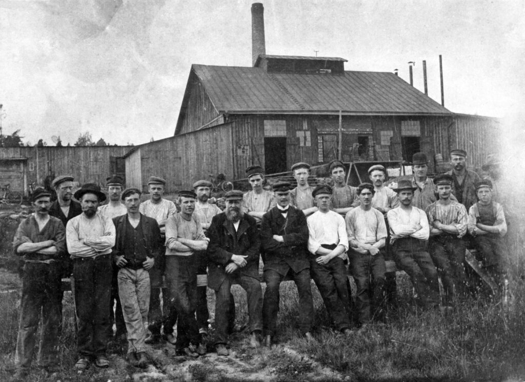 Gruppfotografi, drygt 20 arbetsklädda män står uppställda i två rader framför trähus.