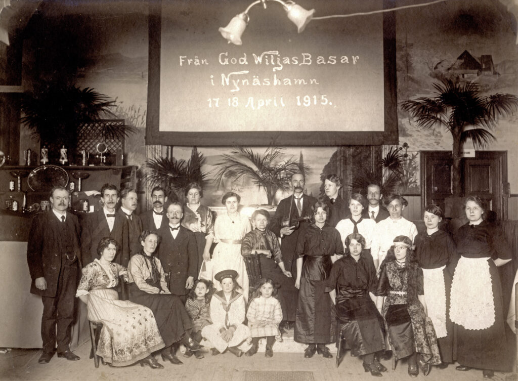 Interiörbild, gruppfoto i logens lokal med sittande och stående finklädda kvinnor, barn och män. I bakgrunden banderoll med texten "Från God Wiljas Basar i Nynäshamn 17 18 april 1915".