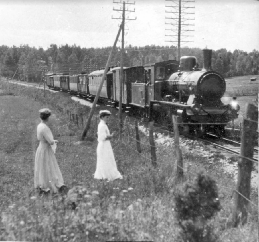 Svartvit bild med två kvinnor i ljusa klänningar som står på en äng. Ett tåg drivet av ett ånglok passerar kvinnorna på nära håll.