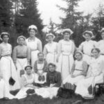 Svartvitt foto med tretton kvinnor som ställt upp sig för fotografering mot en fond av granar.