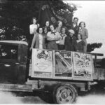 Ett svart-vitt fotografi visar tolv kvinnor som står på flaket till en lastbil. Över dem hänger en fana och på lastbilens sidor sitter affischer.