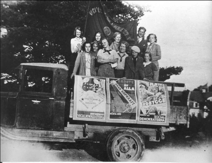 Ett svart-vitt fotografi visar tolv kvinnor som står på flaket till en lastbil. Över dem hänger en fana och på lastbilens sidor sitter affischer.