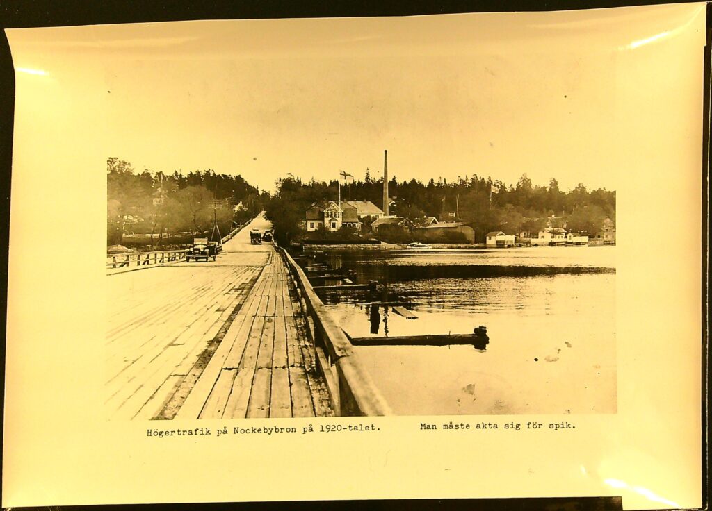 En gammaldags öppen bil kör över en låg träbro. Texten under bilden lyder: Högertrafik på Nockebybron på 1920-talet. Man måste akta sig för spik.
