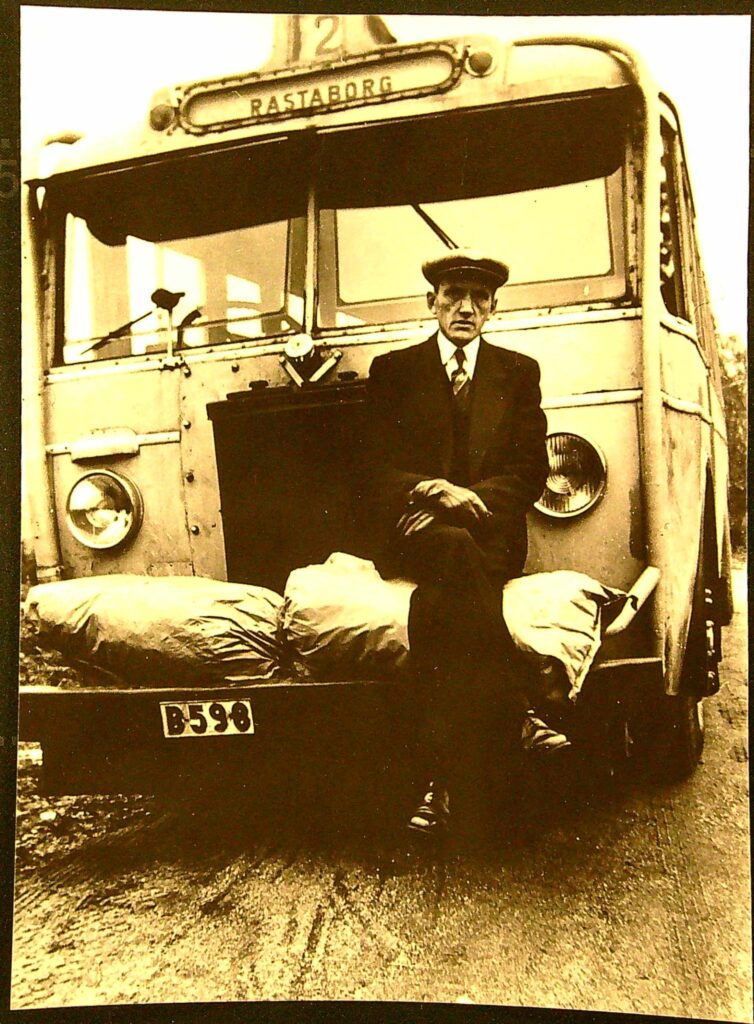 Framsidan av en buss. På kofångaren ligger två stora säckar och där sitter också en man iklädd keps, slips, kostym med väst. Längst upp på bussen står linjens nummer och destination. Vindrutan är tvådelad.