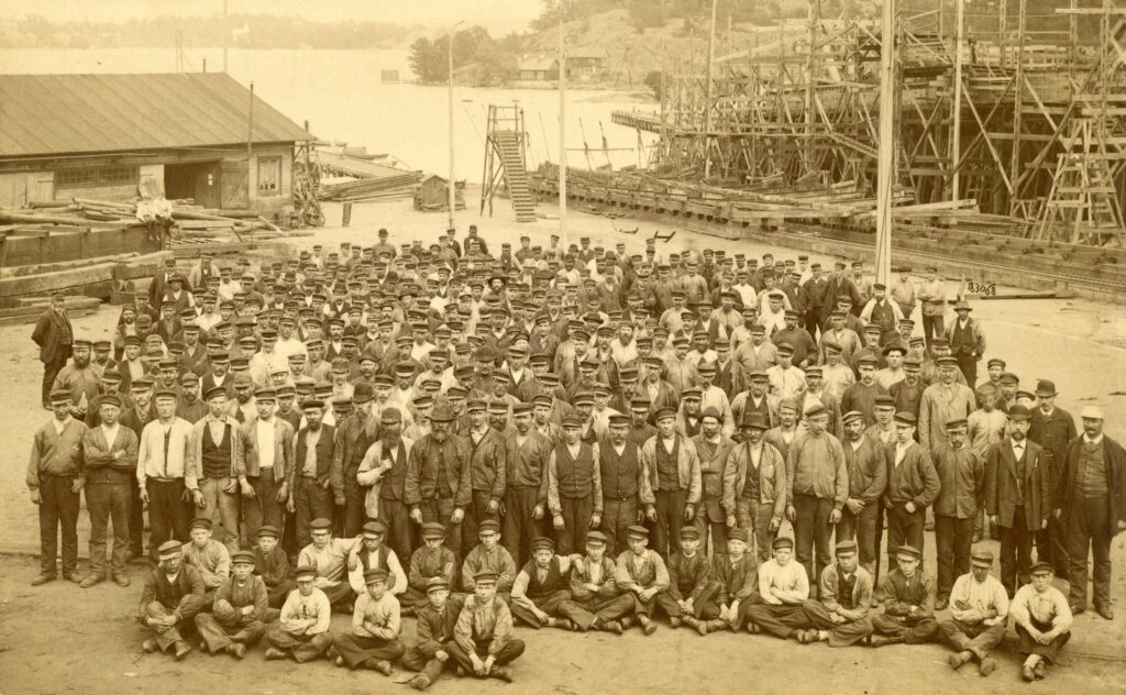 Ett hundratal män står uppställda på skeppsvarvets. Ett tjugotal pojkar sitter i skräddarställning längst fram. I bakgrunden syns byggnadsställningar av trä och havet.