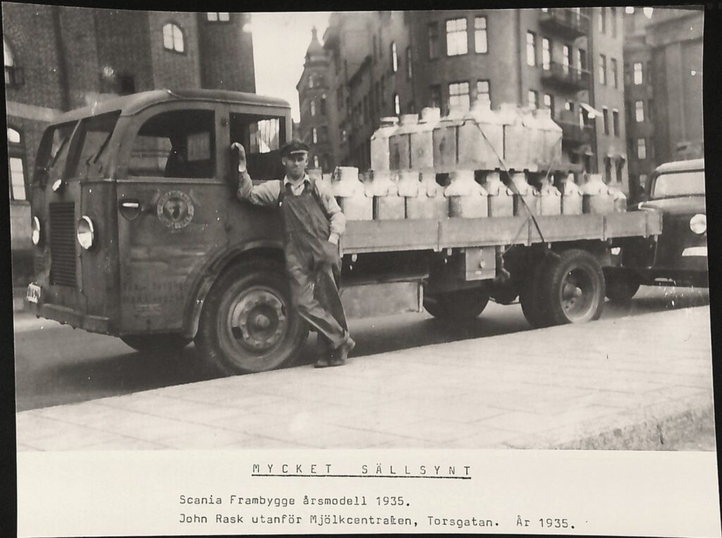 En man i skärmmössa och snickarbyxor står lutat mot en lastbil. På lastbilens flag är ett trettiotal stora mjölkkannor fastspända med remmar. Under bilden står: MYCKET SÄLLSYND, Scania Frambygge årsmodell 1935. John Rask utanför Mjölkcentralen, Torsgatan. År 1935.