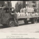 En man i skärmmössa och snickarbyxor står lutat mot en lastbil. På lastbilens flag är ett trettiotal stora mjölkkannor fastspända med remmar. Under bilden står: MYCKET SÄLLSYND, Scania Frambygge årsmodell 1935. John Rask utanför Mjölkcentralen, Torsgatan. År 1935.