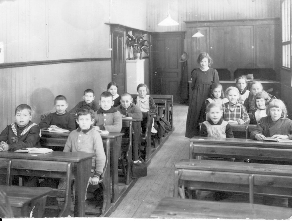 En lärarinna står mellan bänkraderna i ett klassrum. Femton elever sitter i bänkarna.