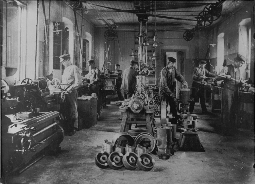 Sju män i skärmmössor arbetar vid olika maskiner i verkstaden.
