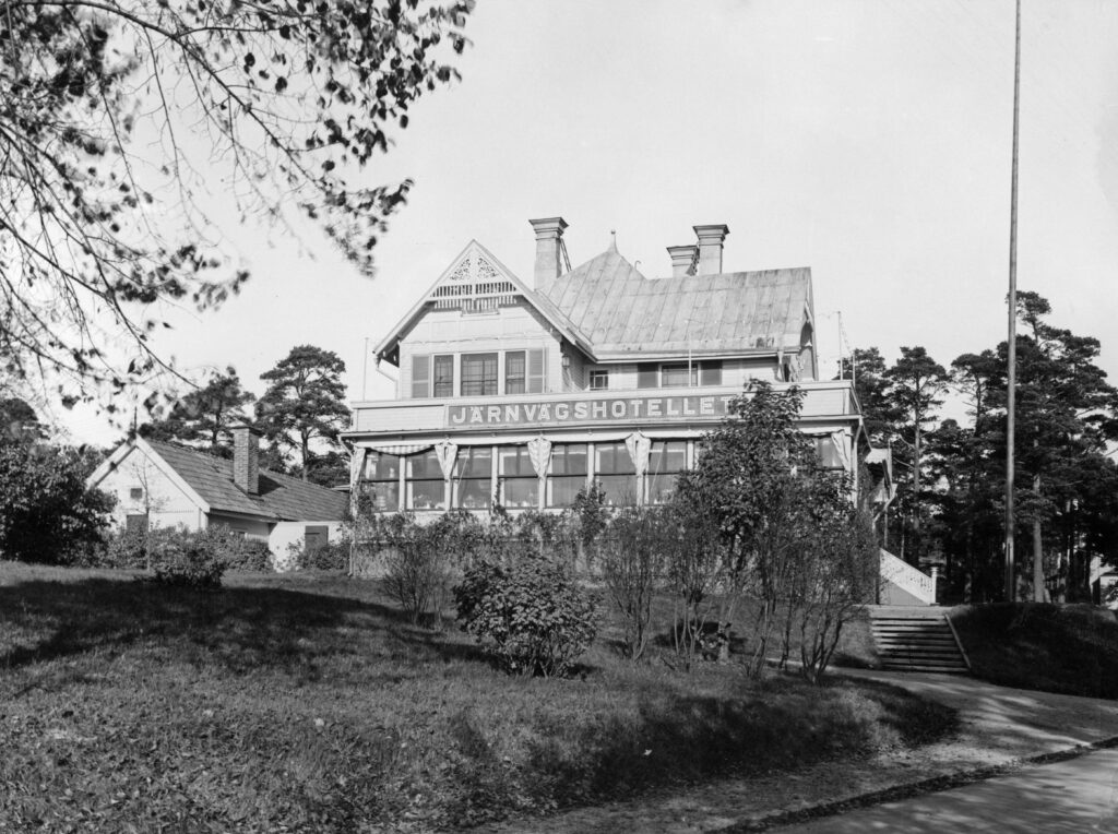 En stor villa med veranda längs hela huset. På fasaden står "Järnvägshotellet". Till vänster ett mindre hus.