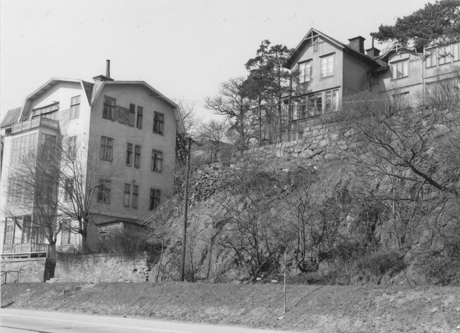 Ett svartvitt foto av två äldre hus. Till höger ett stort hus med fem våningar, till vänster ett mindre hus som ligger uppe på en brant klippa.