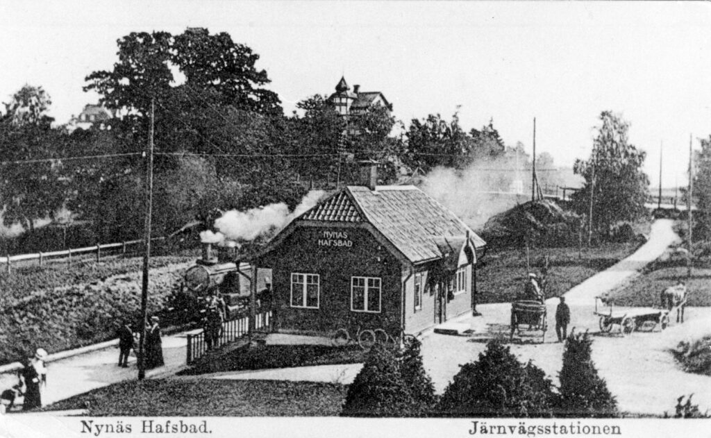 En svartvit bild av det lilla stationshuset där ett rykande ånglok åker på rälsen bortom huset. Bilden är ett vykort och har en text skriven i nederkanten "Nynäs hafsbad. Järnvägsstationen."
