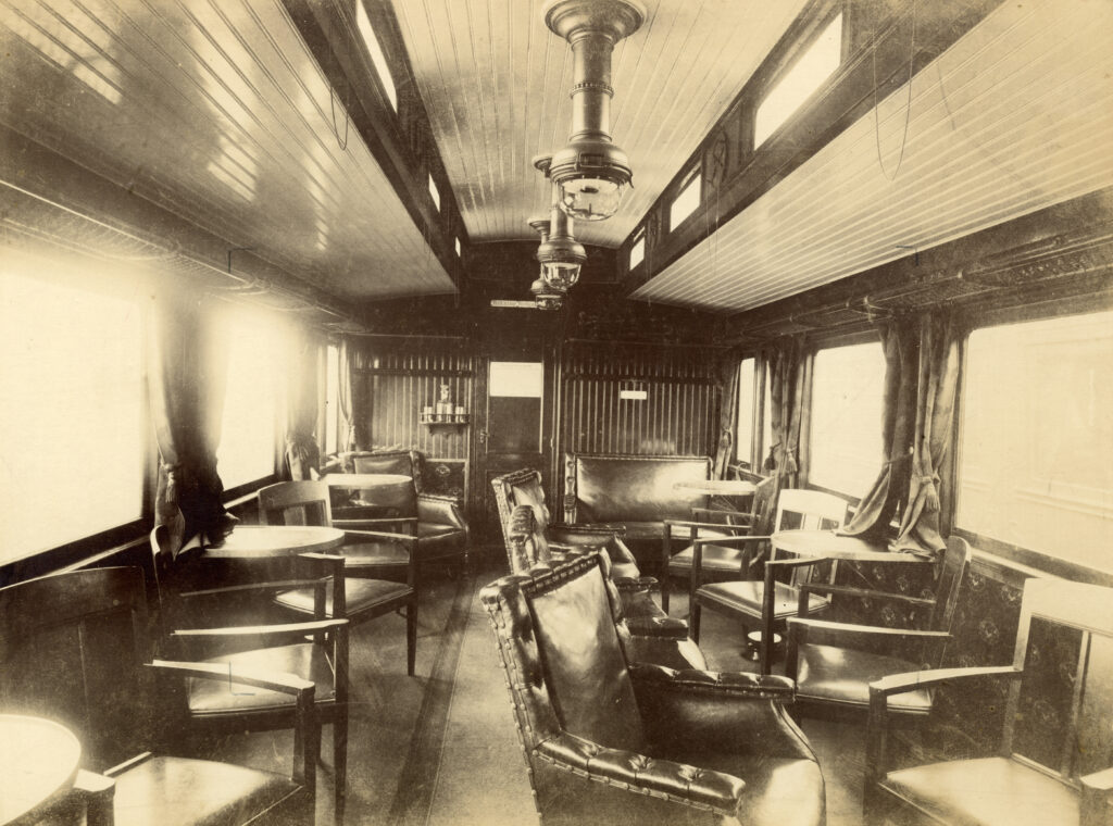 Svartvit interörbild som visar vagnens stolar som står löst på golvet, hatthyllorna, gardinerna och en träpanel på väggen av vagnens kortända.