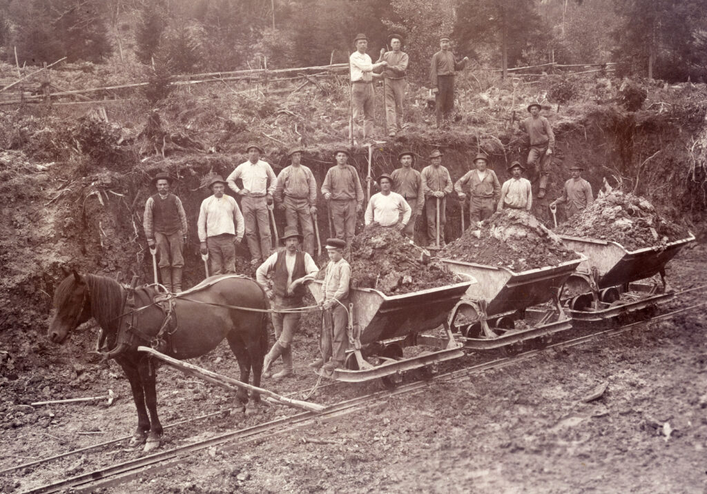 Sjutton rallare står uppställda bakom en häst som spänts för ett litet tåg med tre vagnar fullastade med jord och sten. En av rallarna, en ung pojke står på den främsta vagnen och håller hästens tömmar.