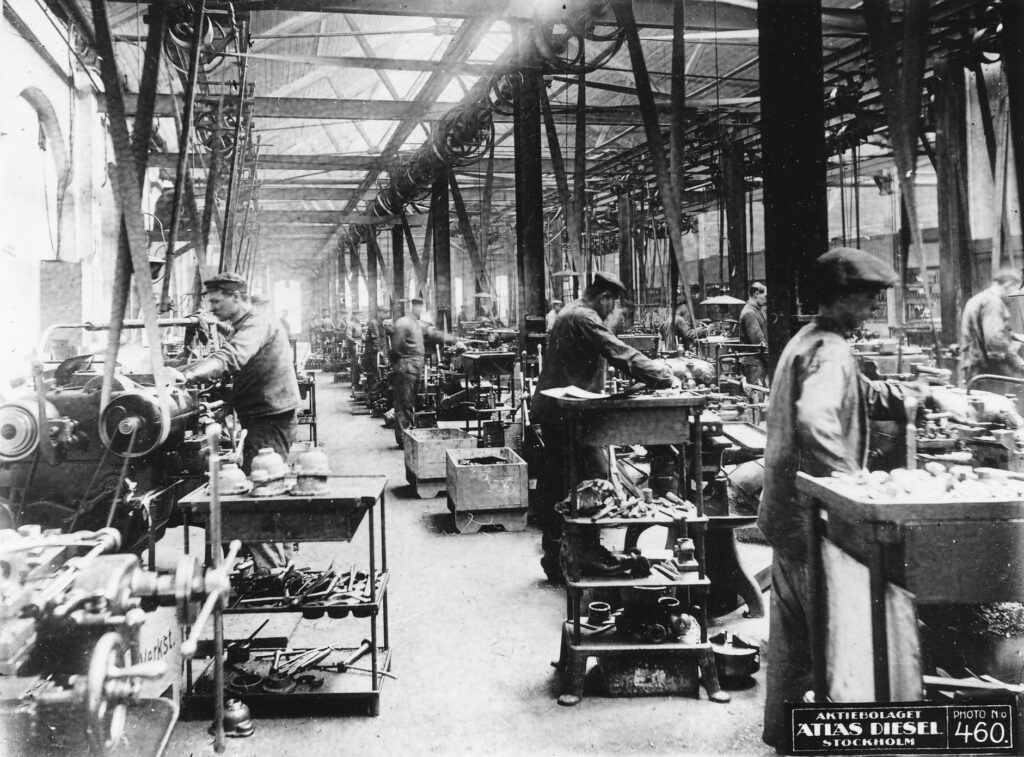 Svartvitt foto av en fabrikshall där män arbetar vid maskiner med drivremmar som går upp i taket.