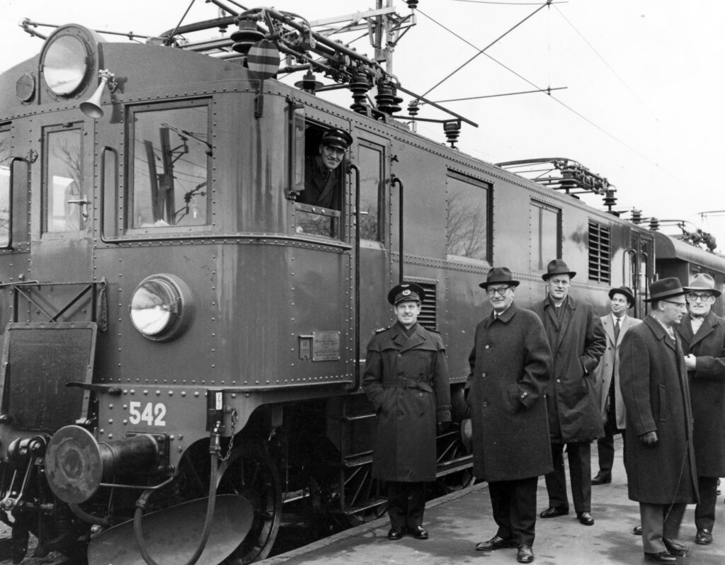 Svartvitt foto av det första eldrivna tåget på Nynäsbanan. En lokförare tittar ut från förarhytten, bredvid tåget står en grupp män.
