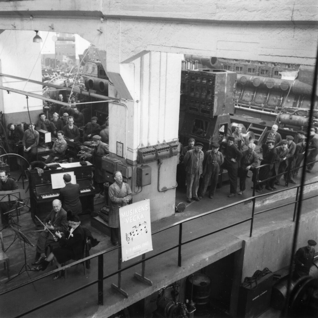Foto taget ovanifrån som visar en sångare och en ackompanjatör vid piano samt två violinister omgivna av arbetare i en fabrik. På ett plakat står "Musikens vecka 2-9 mars 1947".