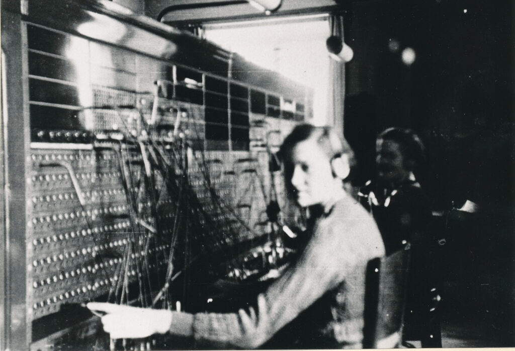 Ett suddigt svartvitt foto visar två kvinnor som sitter med hörlurar framför en vägg med sladdar och kontakter - telefonväxeln.