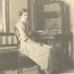 Ett svartvitt fotografi av en kvinna i rutig klänning sitter vid en gammaldags telefonväxel som står i ett rum med småblommiga tapeter.