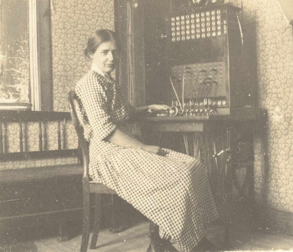 Ett svartvitt fotografi av en kvinna i rutig klänning sitter vid en gammaldags telefonväxel som står i ett rum med småblommiga tapeter.