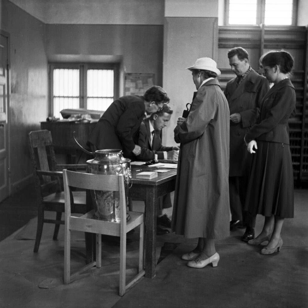 Kvinnor och män i kö till en valurna som står på en stol. En funktionär står böjd över ett bord och antecknar.