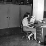 En svartvit bild av en kvinna vid ett skrivbord. På bordet syns ålderdomlig elektronisk apparatur och i bakgrunden syns fyra stora skåpdörrar.