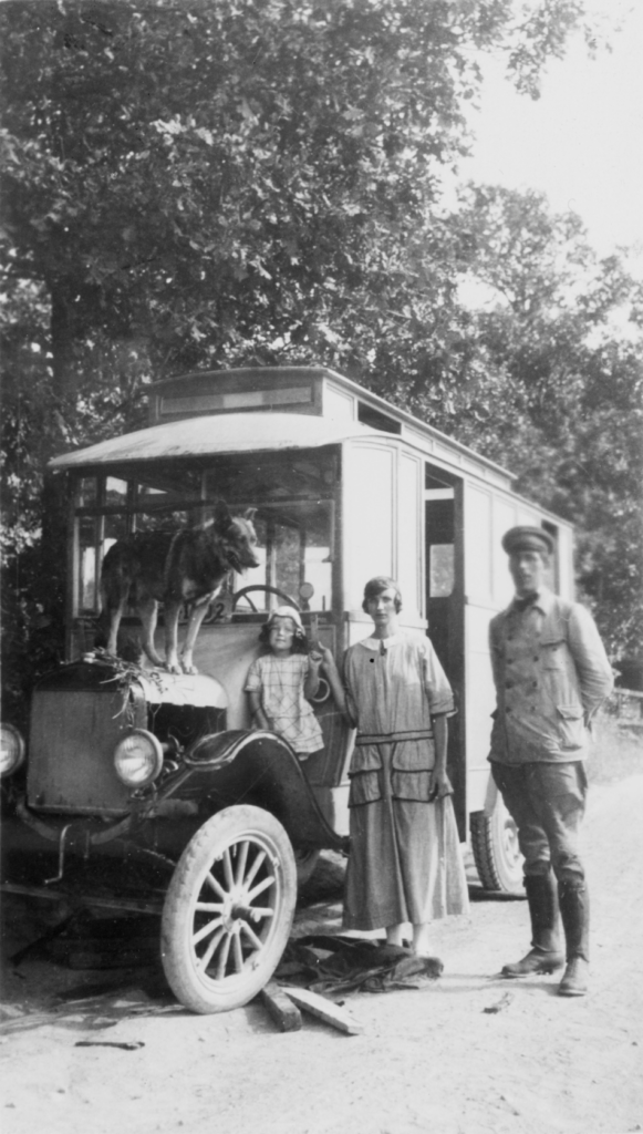 Ett svartvitt foto av en buss. En man och en kvinna står bredvid bussen, på främre stänkskärmen står en liten flicka och på motorhuven en schäferhund.
