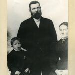 Ett svart-vitt porträtt av familjen där pappan i stort skägg står upp flankerad av mamman och sonen som sitter på var sin sida.