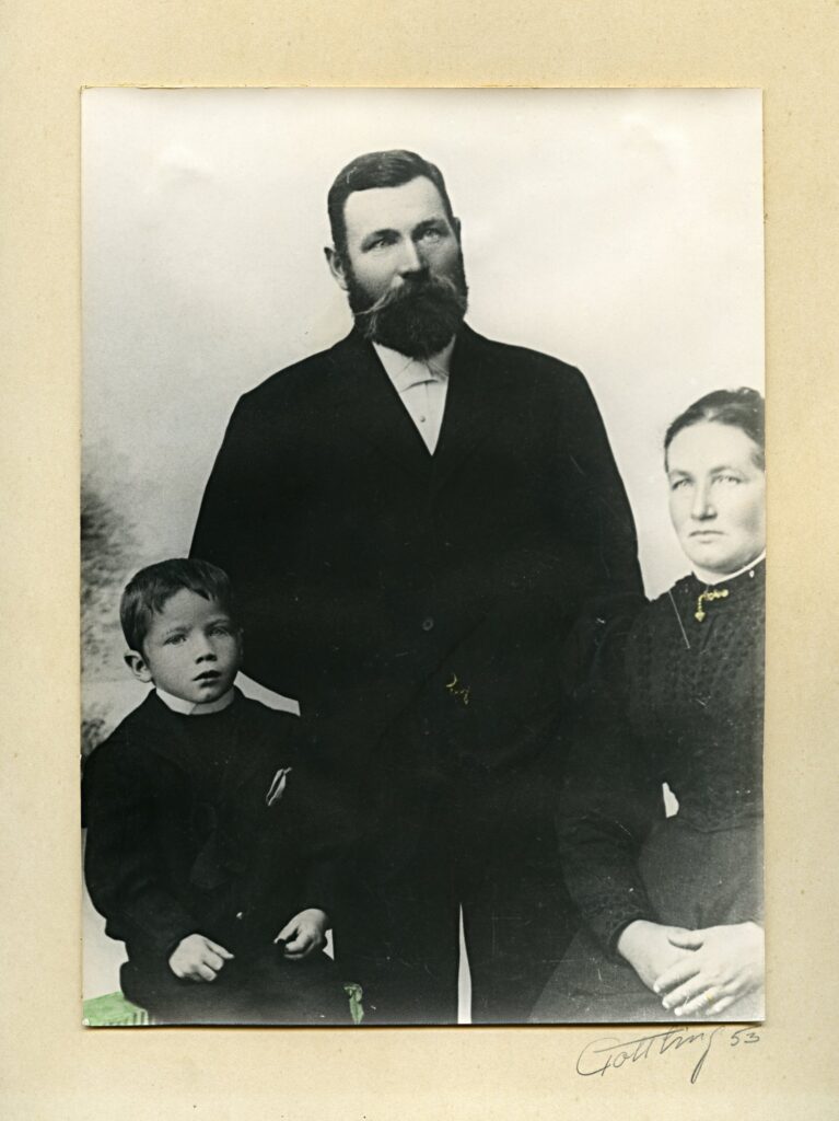 Ett svart-vitt porträtt av familjen där pappan i stort skägg står upp flankerad av mamman och sonen som sitter på var sin sida.