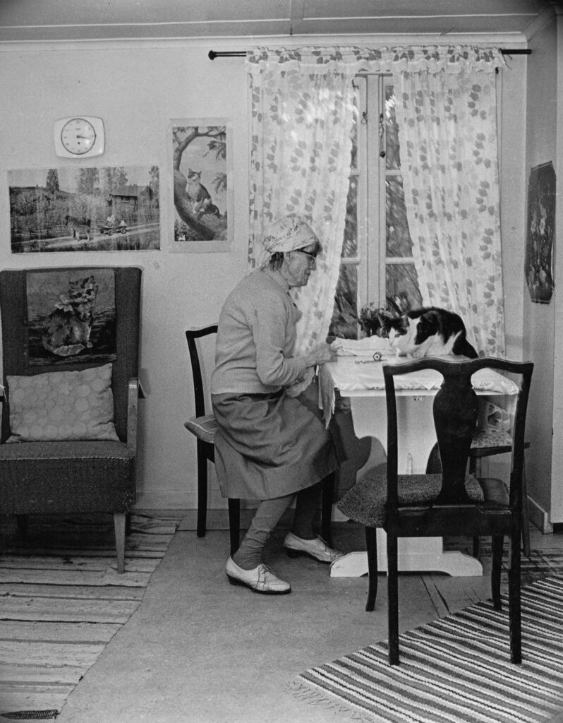 Ett svartvitt foto av en kvinna i huckle som sitter vid ett bord med en katt.
