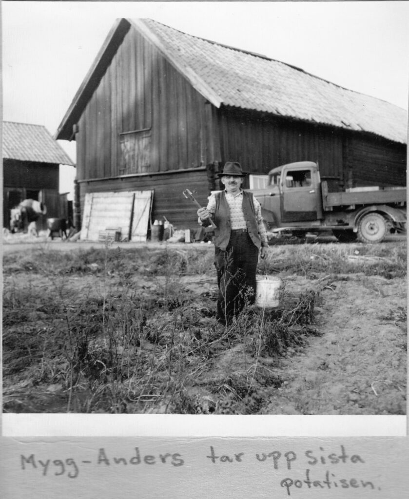 Svartvitt foto av en man i hatt, väst och mustasch. Han håller en hink i ena handen och en hacka i den andra. I bakgrunden syns ladugård och en ålderdomlig lastbil.