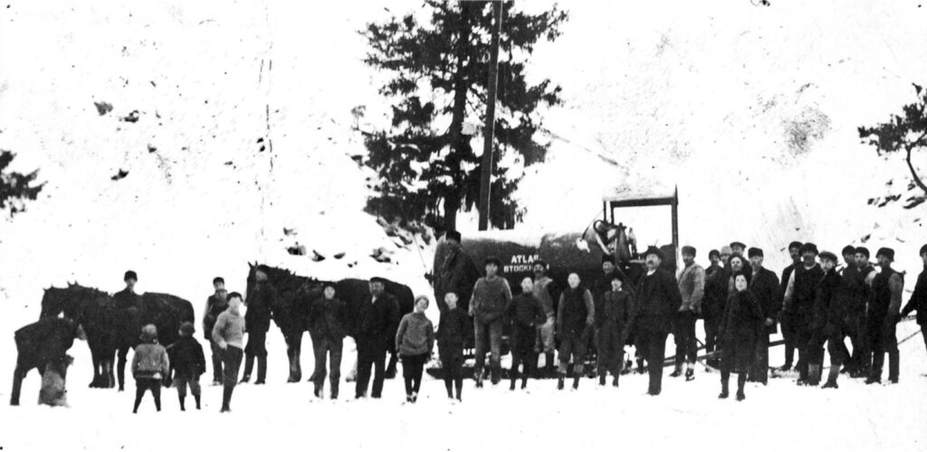 Ett svartvitt foto med ca 25 personer framför ett lok och två hästar.