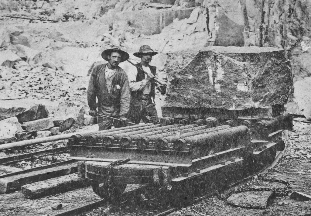 foto av två män i hatt och mustasch vid en vagn på räls. På vagnen står ett stenblock.