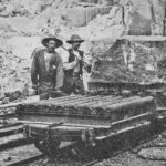 foto av två män i hatt och mustasch vid en vagn på räls. På vagnen står ett stenblock.