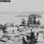 Ett svartvitt foto av några hus vid vattnet omgivna av högar av trä och virke.