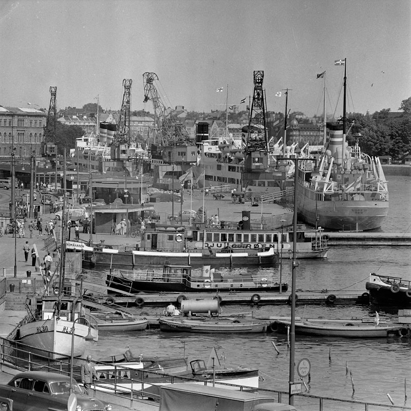 Ett svartivtt foto av båtar vid Skeppsbron. Fyra stora lyftkranar finns på kajen och vattnet är fullt av båtar.