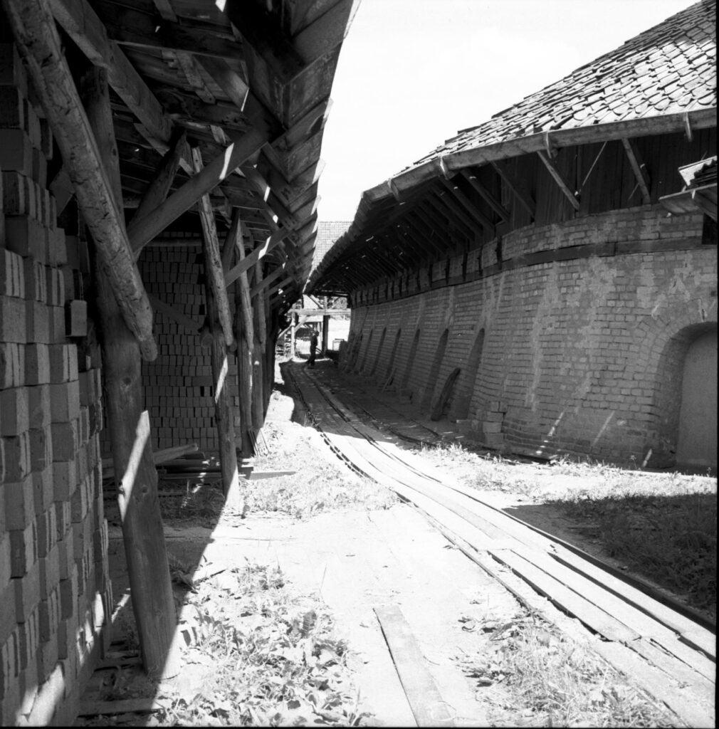 Svartvitt foto av en järnvägsräls som går mellan två lador. Den en är murad av tegel, den andra har en öppen vägg där stora högar av tegel syns.