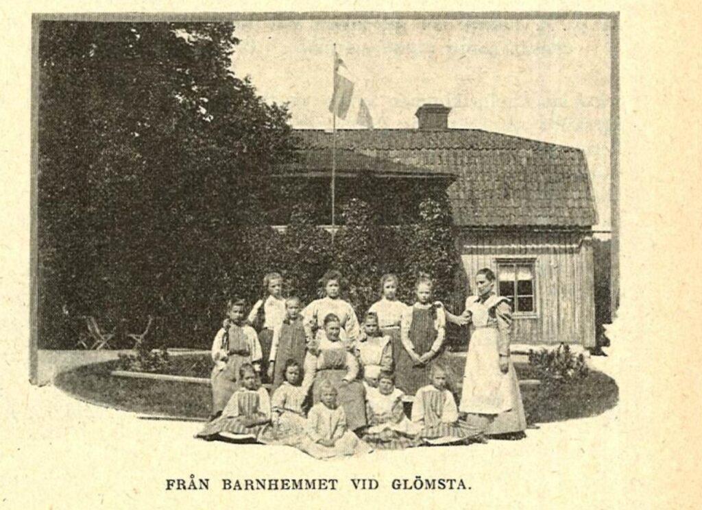 Tretton flickor står uppställda framför Glömsta gård tillsammans med sin föreståndarinna. Texten under bilden lyder: Från barnhemmet vid Glömsta.