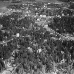 Ett svartvitt flygfoto med villor som sticker fram i skogen. Längre bort syns öppnafält och järnväg.