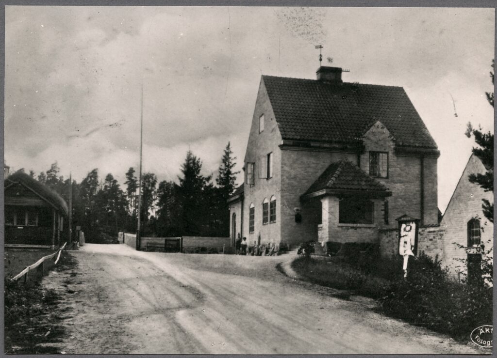 Svartvitt foto av stationshuset i putsat tegel till höger om vägen och en liten stuga med grästak till vänster.