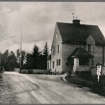 Svartvitt foto av stationshuset i putsat tegel till höger om vägen och en liten stuga med grästak till vänster.