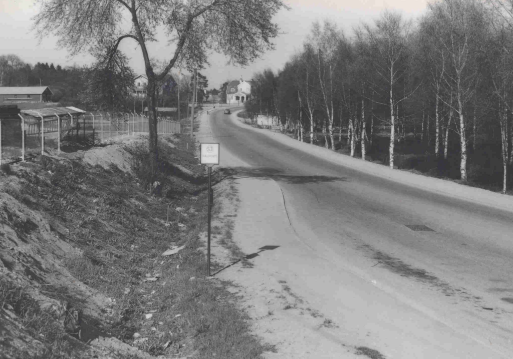 Ett svartvitt foto av en landsväg som leder upp för en backe till några hus. Mitt i bilden står en skylt.