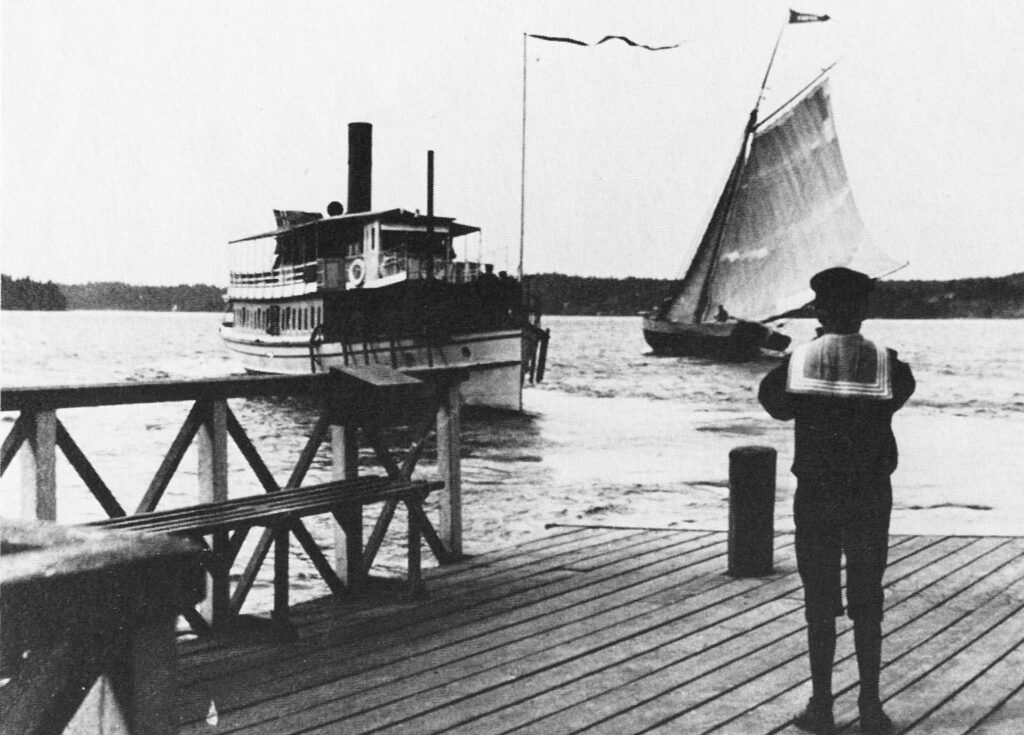 En pojke i sjömanskostym står med ryggen mot fotografen och tittar på en ångbåt och en segelbåt med hissat segel.