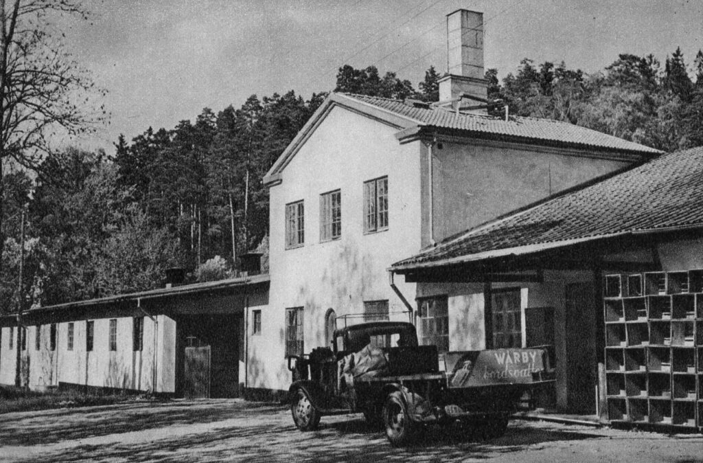 Svartvitt foto av en fabriksbyggnad med en ålderdomlig lastbil som har en skylt på flaket. Texten på skylten lyder Wårby bordsvatten.