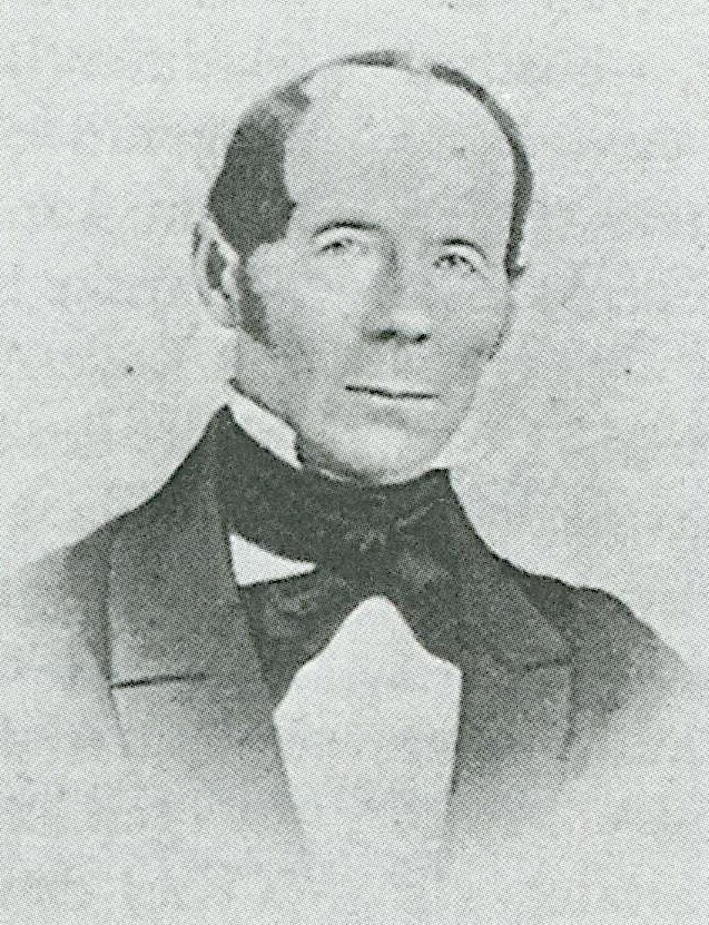 Ett svartvitt porträttfoto av en man med handknuten mörk fluga, kavaj, polisonger och kal hjässa.