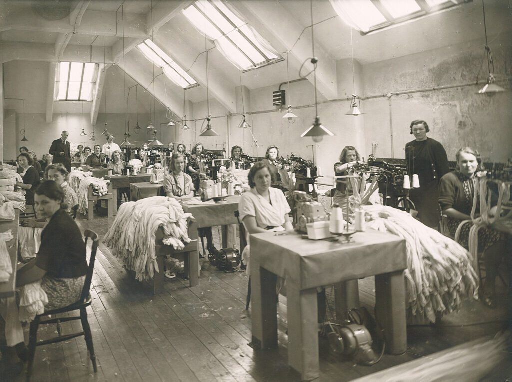 Kvinnor sitter vid symaskiner i ett stort rum med takfönster.